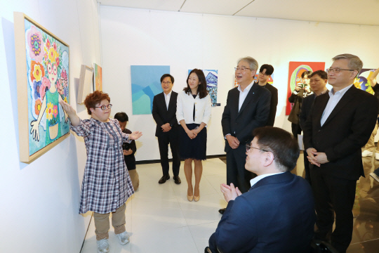 우리금융미래재단이 오스트리아 빈에서 주최하는 '2023 한-EU 발달장애 미술가 해외교류전'에 앞서 지난 8일 서울 중구 우리금융 본사에서 출정식과 국내 전시회 개회식을 개최했다. 참여 작가가 우리금융미래재단 임종룡(뒷줄 오른쪽 두번째) 이사장(우리금융 회장) 등이 참석한 가운데 우리금융 본사에서 열린 국내 전시회에서 작품을 설명하고 있다.  우리은행 제공